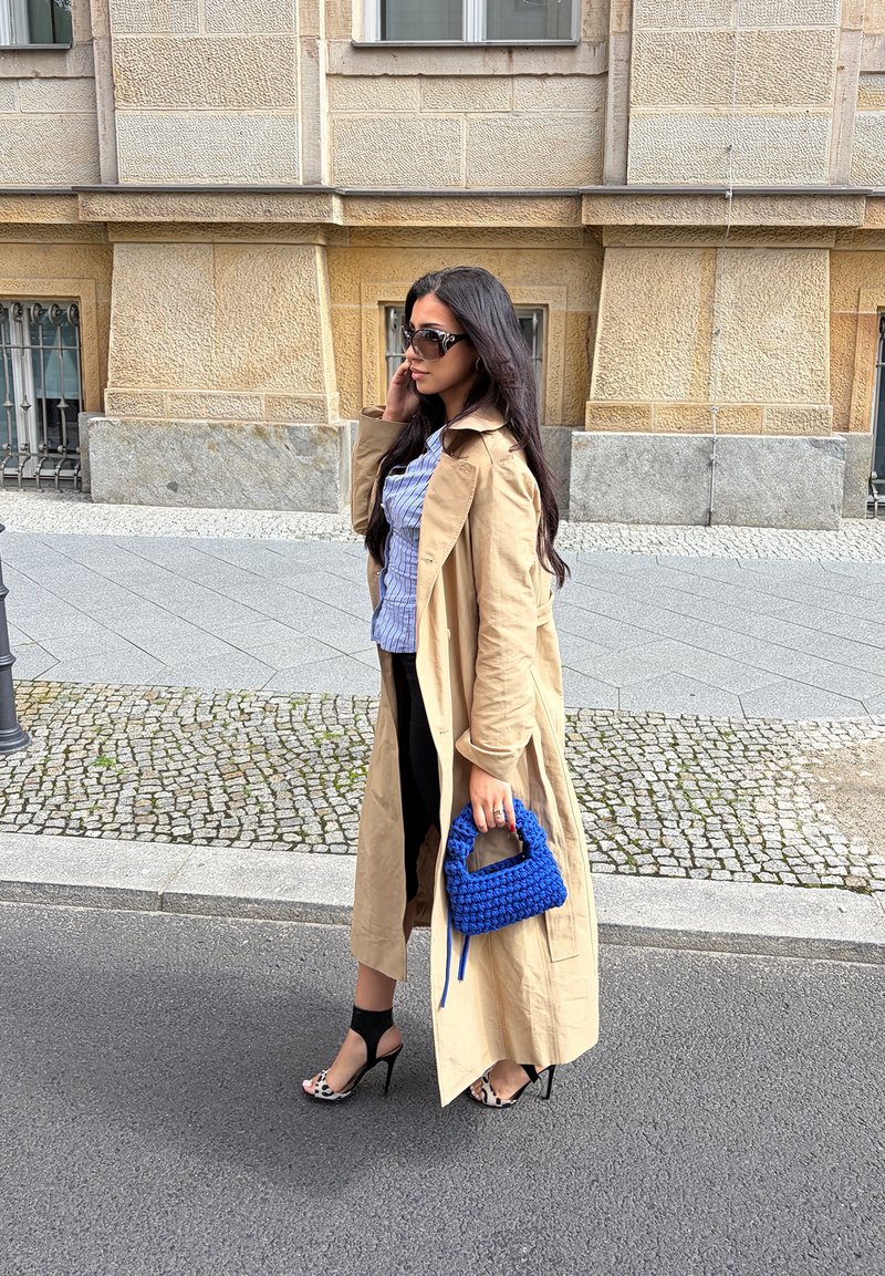 Kvinna med långt mörkt hår, bär solglasögon, beige trenchcoat, svart kjol och leopardmönstrade klackar, håller en blå handväska på en stadsgata.