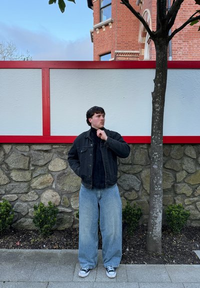 Joven con chaqueta negra y pantalones vaqueros anchos, de pie junto a un muro de piedra con una verja roja y blanca y un árbol, en un entorno urbano al aire libre.