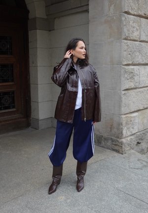 Mujer de pie al aire libre junto a una pared de piedra y una puerta de madera, vestida con una chaqueta de cuero marrón, pantalones deportivos azules con rayas blancas y botas marrones.
