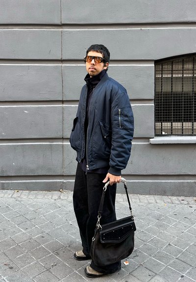 Hombre con chaqueta oscura y gafas de sol, de pie en una calle pavimentada, sosteniendo un bolso negro frente a una pared gris con una ventana con rejas.