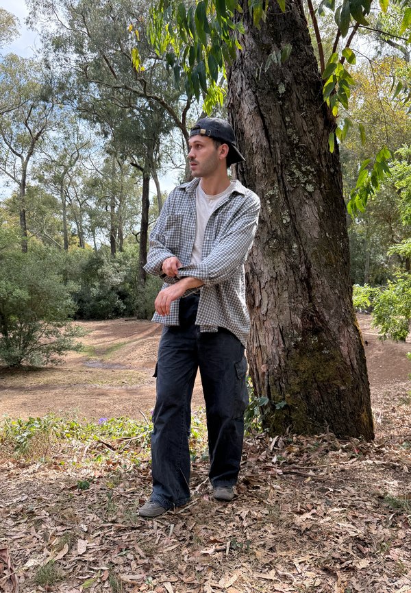 Un hombre con gorra y camisa de cuadros se arregla la manga junto a un gran árbol en un parque arbolado.