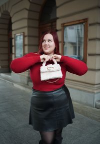 Λευκή φουντωτή τσάντα με χρυσή αλυσίδα, κρατημένη από ένα άτομο που φοράει κόκκινο πουλόβερ. Η μαύρη δερμάτινη φούστα προσθέτει αντίθεση στην εμφάνιση. Αστικός φόντος.