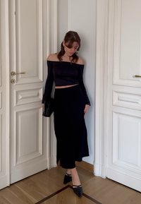 Jeune femme portant un haut noir à épaules dénudées et une longue jupe, debout à l'intérieur entre deux portes blanches, tenant une pochette noire.