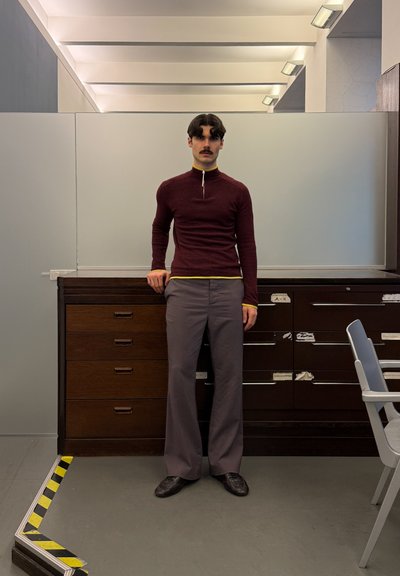 Hombre con cabello oscuro y bigote, vestido con un suéter burdeos con cremallera y pantalones grises, de pie en el interior junto a armarios de madera y una silla.