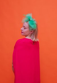 Φωτεινό ροζ κάπα με λεπτομέρεια σε σχήμα κλειδώματος, συνδυασμένο με μια μεγάλη πράσινη τulle στεφάνη που διαθέτει ένα μαργαρίτη, σε πορτοκαλί φόντο.