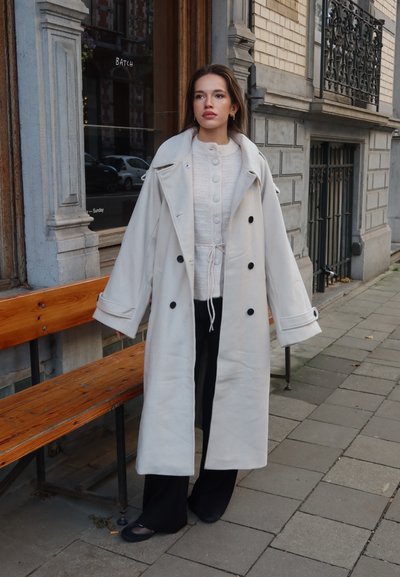 Abrigo trinchado color crema con grandes botones negros, superpuesto a una blusa de punto blanca y pantalones de pernera ancha negros. Tejido suave, diseño oversize.