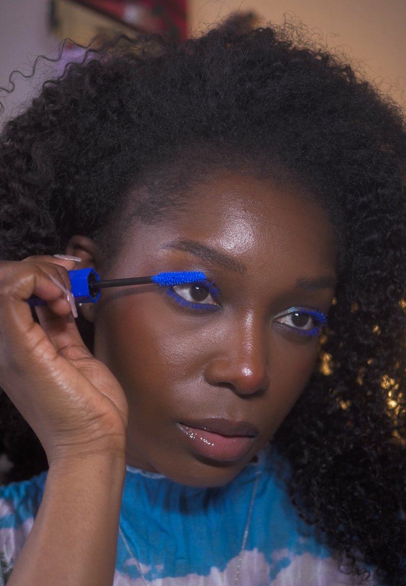 Personne appliquant du mascara bleu vif sur les cils supérieurs, portant un haut bleu tie-dye, avec des cheveux noirs bouclés et une expression concentrée.