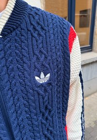Tmavomodrý pletený sveter s vrkočovým vzorom, vrátane bieleho vyšívaného loga Adidas. Rukávy majú kontrastné textúry a farby.