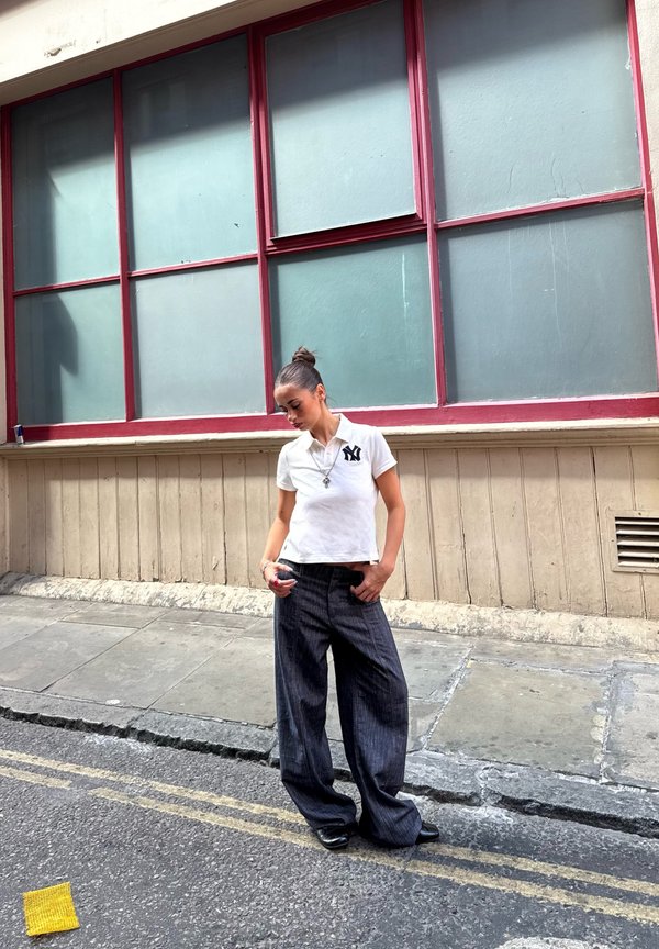 Giovane donna con i capelli raccolti in uno chignon indossa una polo bianca e pantaloni larghi scuri, in piedi su una strada della città vicino a un edificio con grandi finestre incorniciate di rosso.