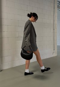 Χοντρή καρό oversized σακακιού σε γκρι απόχρωση, μπλε τζιν σορτς, μαύρη πλεκτή τσάντα, λευκές κάλτσες και μαύρα παπούτσια με λουρί. Φόντο με τσιμεντένιο τοίχο.