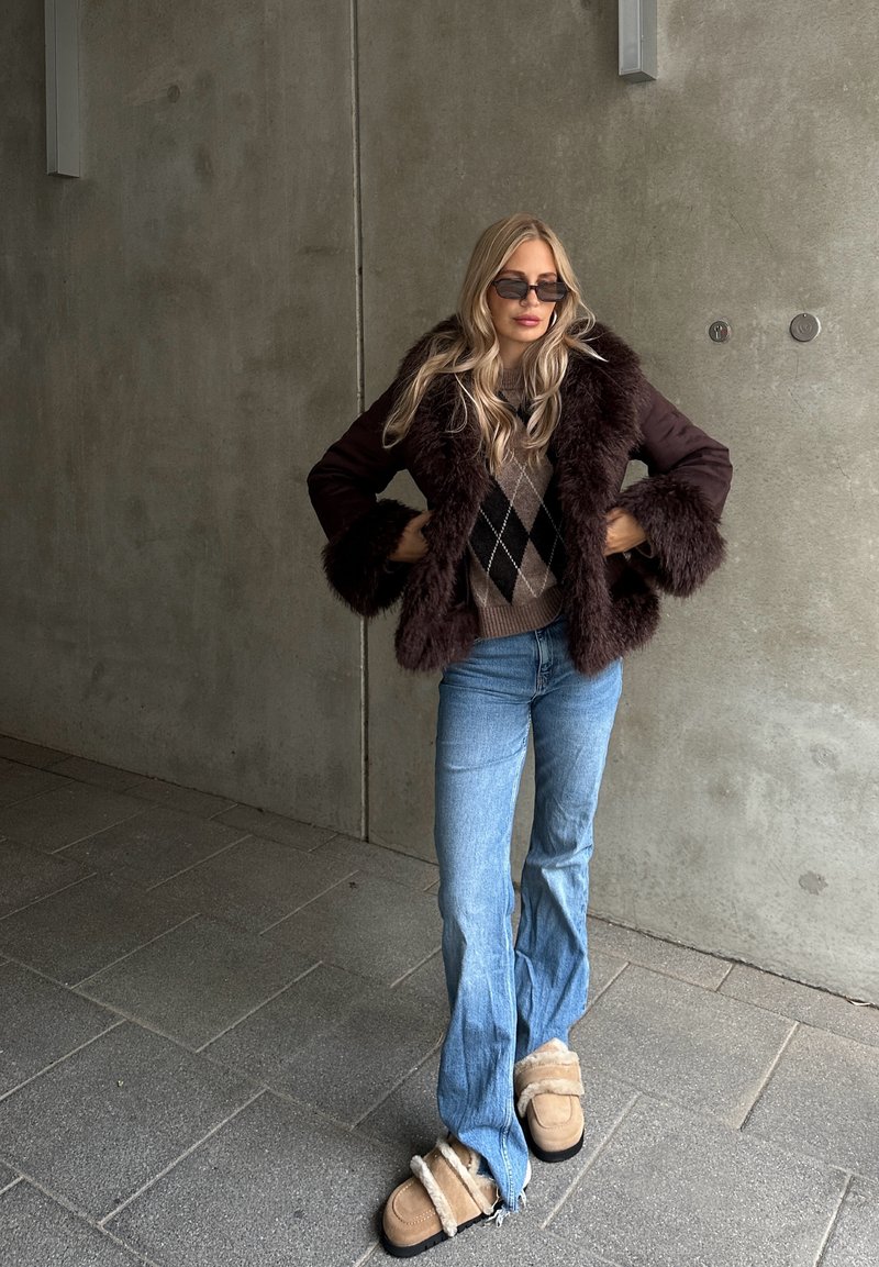 Brauner Kunstpelzmantel mit passenden Manschetten, Argyle-Muster-Pullover, blaue Schlagjeans und beige Shearling-Pantoletten.