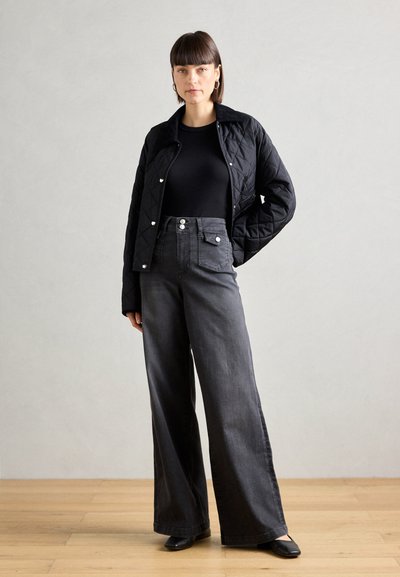 Chaqueta negra acolchada, top negro ajustado, jeans de pierna ancha de tiro alto en gris oscuro y zapatos negros sobre un suelo de madera contra un fondo neutral.