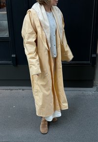 Langer, blassgelber Mantel aus leichtem Material, mit einer großen Kapuze und übergroßen Ärmeln. Getragen über einem grauen Pullover und einem weißen Rock.