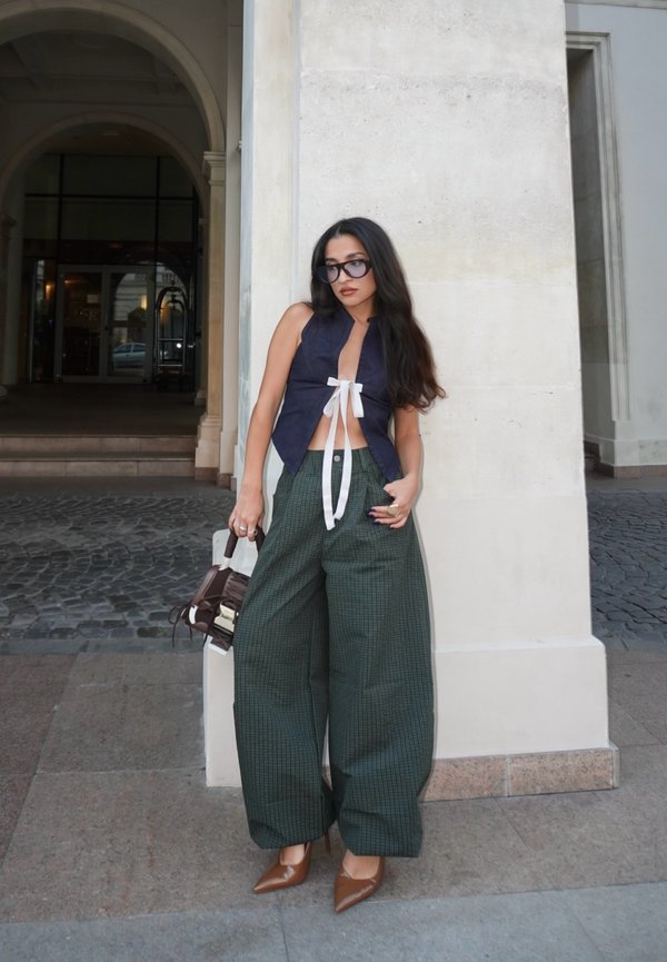 Mujer con pantalones verdes anchos, top sin mangas azul marino con lazos blancos, tacones marrones y gafas de sol oscuras, de pie junto a una columna de piedra al aire libre.
