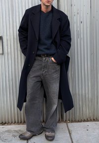 Long manteau bleu marine, pull bleu foncé, jean gris oversized et chaussures noires. Texture lisse et coutures visibles. Fond en béton.