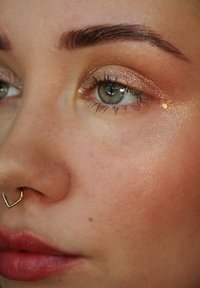 Nærbilde av et ansikt med lys brun hud, grønne øyne og fremtredende kinnben. Ansiktet har glitrende øyemakeup og en hjerteformet septumring i gull.