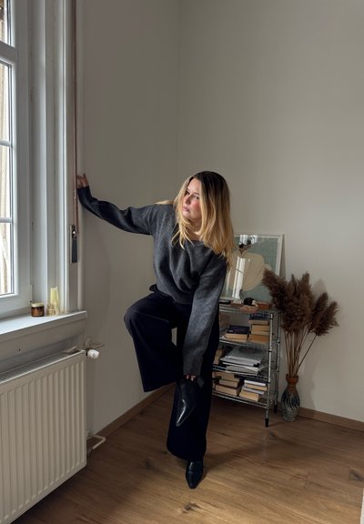 Mujer con suéter gris oscuro y pantalones negros ajustándose una bota negra en un tobillo junto a una ventana en una habitación minimalista con libros y plantas secas.
