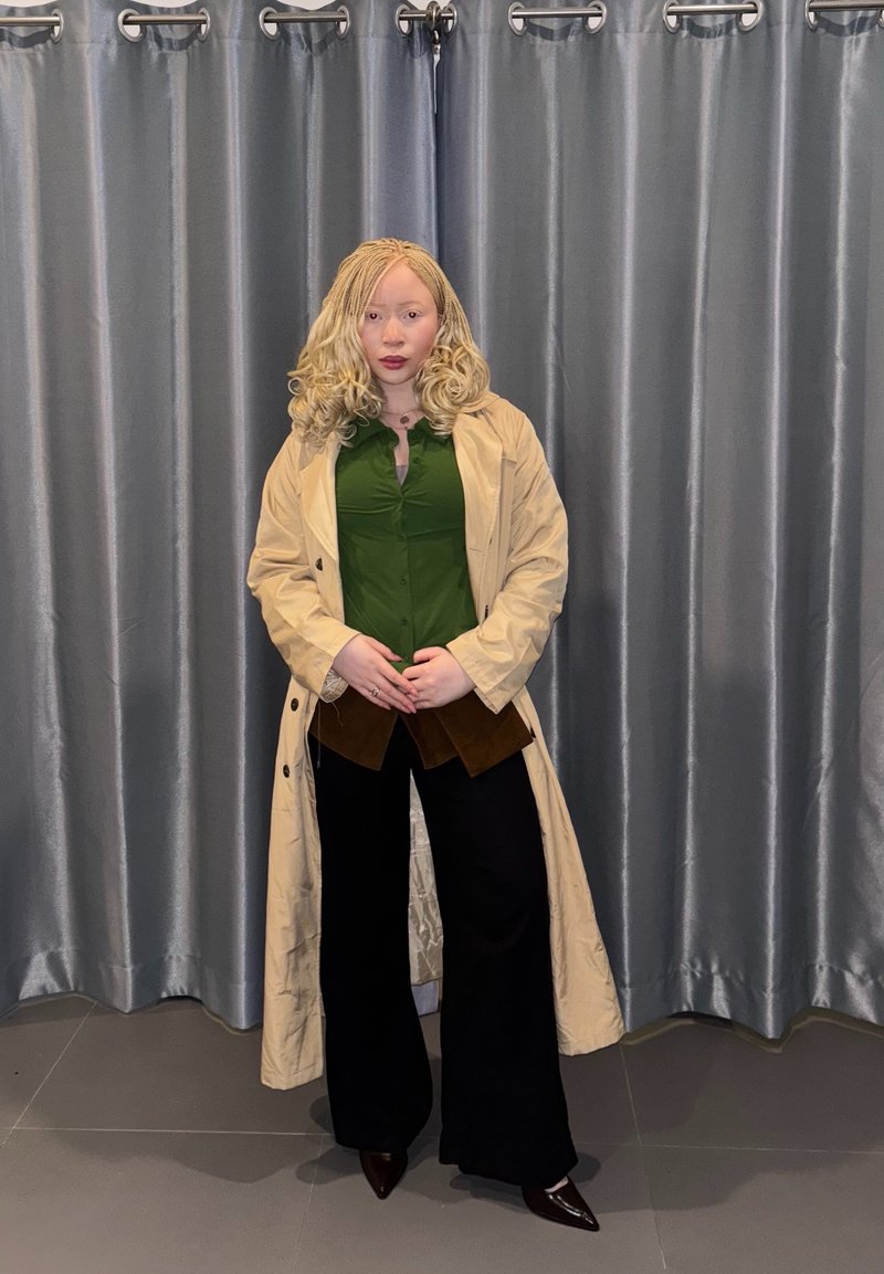 Henkilö, jolla on vaaleat kiharat hiukset, pukeutunut beigeen pitkässä takissa, vihreään paitaan, mustiin leveälahkeisiin housuihin ja ruskeisiin teräväkärkisiin kenkiin, seisoo harmaiden verhojen edessä.