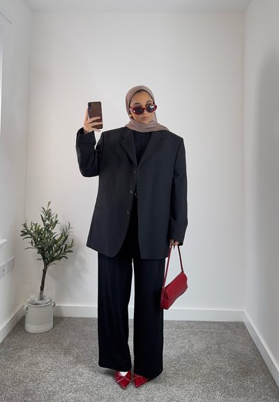 Blazer negro de gran tamaño sobre una blusa negra y pantalones de pierna ancha, combinados con gafas de sol rojas y un pequeño bolso rojo, ante un fondo neutro.