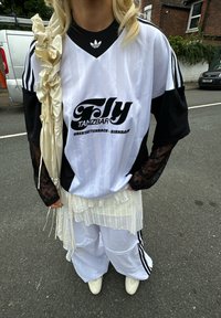 Hvit og svart stripete sportsjersey med logo, kombinert med oversized hvite bukser, volangdetalj i hvitt og blonde lange ermer i svart. Hvite sko.