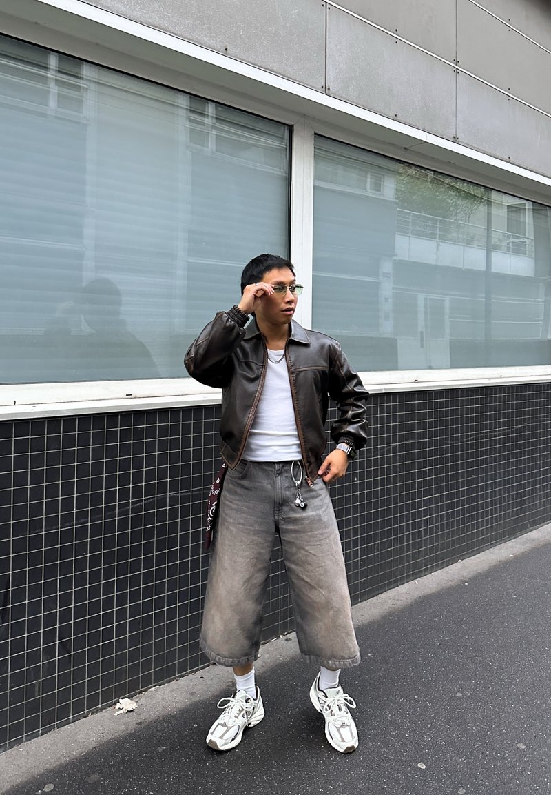 Casaco bomber castanho de couro, t-shirt branca, jeans cinza claro cropped e ténis brancos. Os acessórios incluem óculos transparentes e uma bandana.