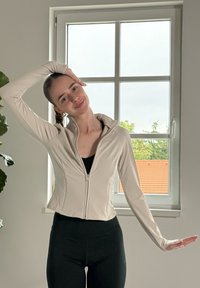 Svetlo bež jakna na zadrgo s prilegajočim dizajnom in visokim ovratnikom, v kombinaciji s črnimi leggins. Tkanina se zdi gladka in raztegljiva.