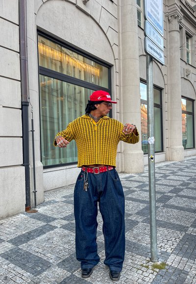 Suéter de punto a rayas amarillas y negras con un escote pronunciado; pantalones de mezclilla azul flojos; cinturón rojo con tachuelas; zapatos negros.