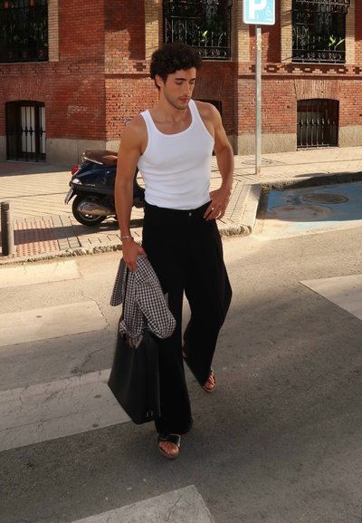 Pantalones de pierna ancha negros, camiseta sin mangas blanca, bolso de mano negro y sandalias marrones. Lleva una camisa a cuadros gris y blanca. Entorno urbano elegante.
