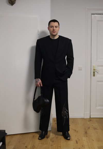 Hombre con blazer negro y pantalones de gran tamaño con diseños de llamas, sosteniendo un bolso negro, de pie en el interior sobre un suelo de madera.