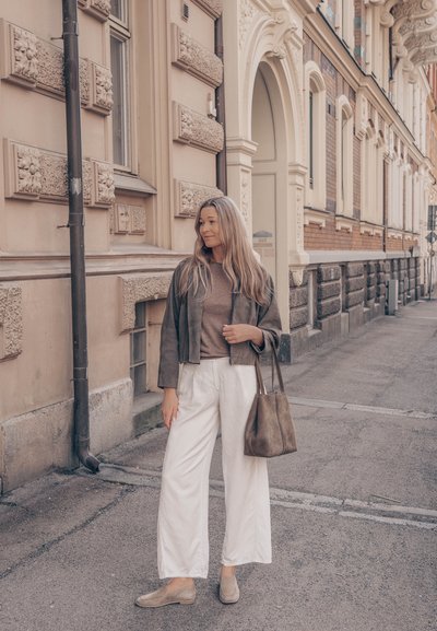 Mujer con cabello largo, vestida con chaqueta marrón, camiseta beige, pantalones blancos y mocasines, sosteniendo un bolso tote marrón, de pie en una calle de la ciudad.