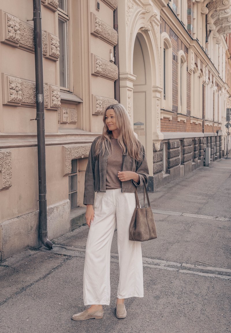 Femme aux longs cheveux portant une veste marron, un haut beige, un pantalon blanc et des mocassins, tenant un sac cabas marron, debout dans une rue de la ville.