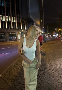 Mudel valges ilma varrukateta tipis ja beežis nööriga seelikus. Linnaline öömaastik, kus on kiviparket ja linnatuled taustal.