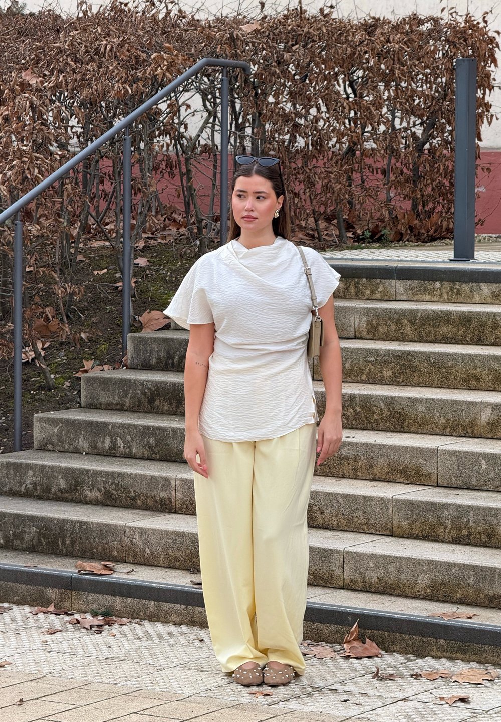 Λευκή υφασμάτινη μπλούζα με φουσκωτά μανίκια, συνδυασμένη με χαλαρές κίτρινες παντελόνες. Καφέ επίπεδες παπούτσια με διακοσμητικά. Υπαίθριο περιβάλλον με σκαλοπάτια.