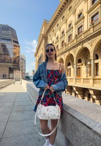 Λουλουδάτο φόρεμα με κόκκινες, ροζ και μπλε παραστάσεις, συνδυασμένο με τζιν jacket. Κρατάει μια λευκή τσάντα με λουλουδάτες λεπτομέρειες και μακρύ λουρί.