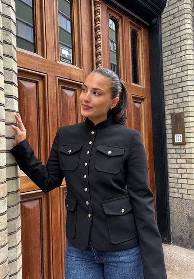 Chaqueta negra entallada con cuello alto, cuatro bolsillos con solapa y botones dorados. Lleva puesta sobre jeans de mezclilla azul. Acabado de tela texturizada.