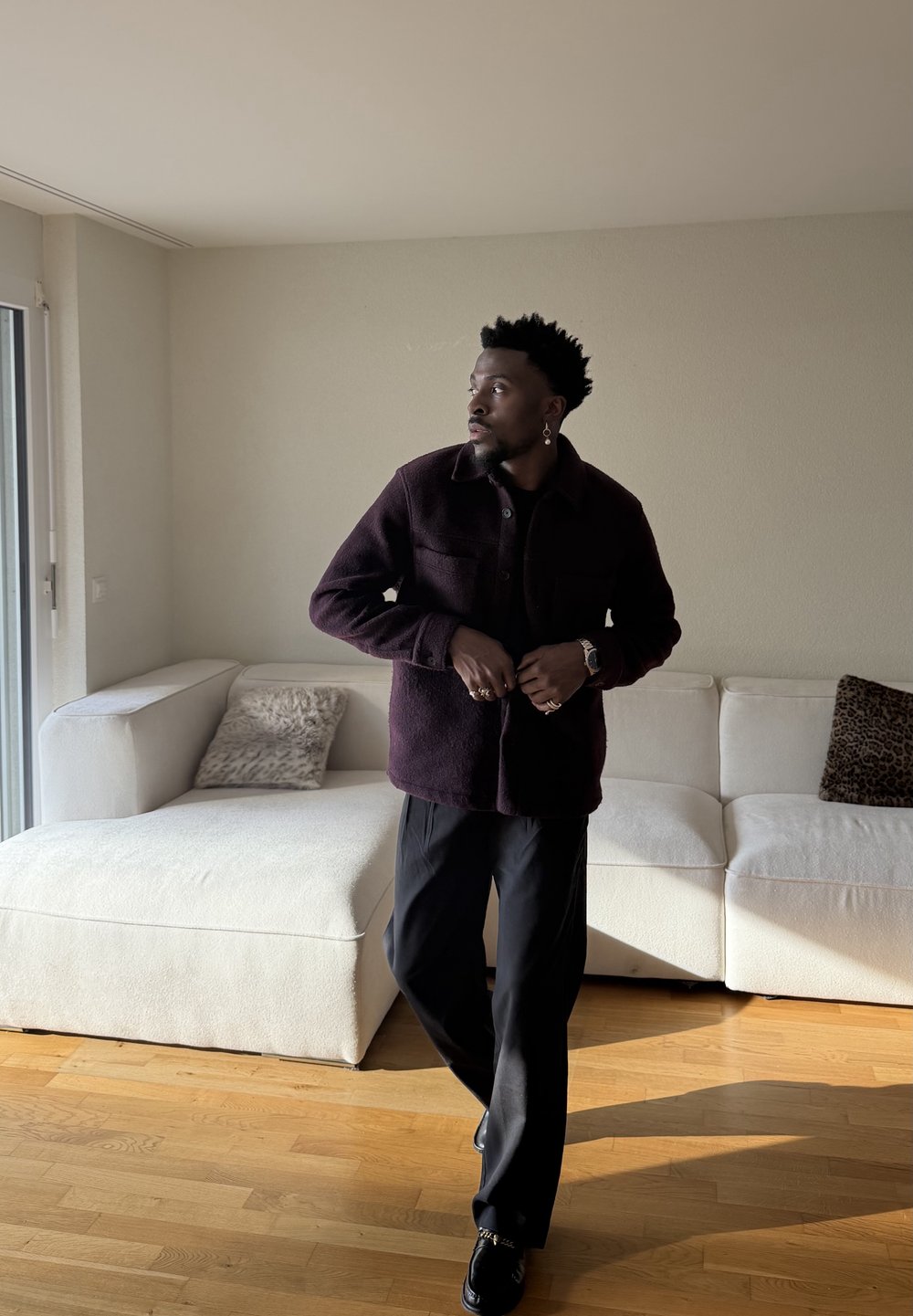 Un homme portant une veste et un pantalon foncés se tient dans un salon baigné de lumière avec un canapé d'angle blanc et des coussins décoratifs.