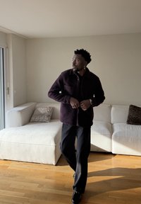 Un homme portant une veste et un pantalon foncés se tient dans un salon baigné de lumière avec un canapé d'angle blanc et des coussins décoratifs.