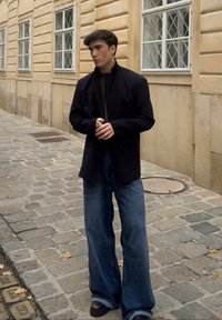 En person står på brostein iført en mørk blazer over en svart rullekragegenser og vide blå jeans, fremhevet av svarte platåsko.