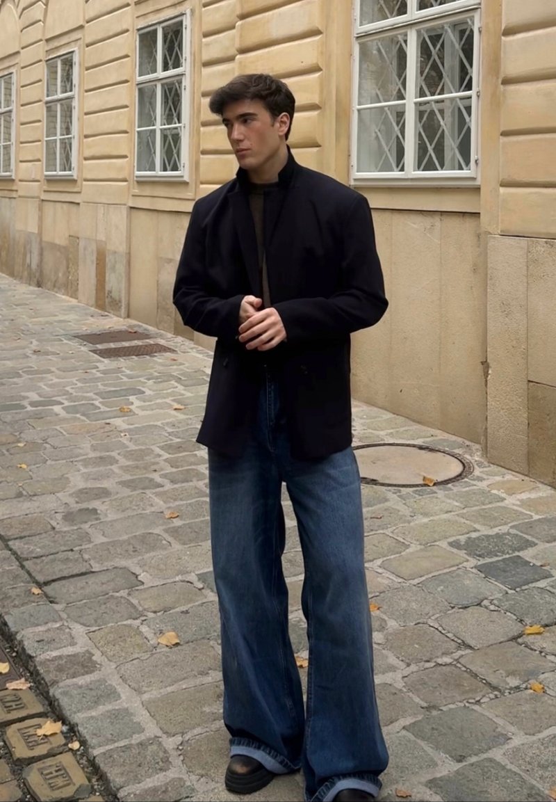 Asmuo stovi ant akmeninio grindinio, dėvi tamsų švarką ant juodos golfos ir plačių mėlynų džinsinių kelnių, akcentuotas juodais platforminiais batais.