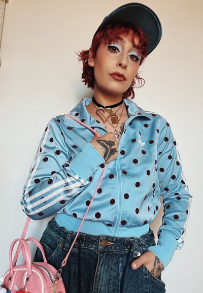 Persona con cabello rojo que lleva una chaqueta azul con lunares y una gorra negra, sosteniendo una bolsa rosa, mostrando tatuajes de mariposas en las manos y collares en el pecho.