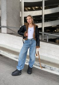 Chaqueta de piel negra corta con forro de piel sintética marrón, camiseta sin mangas blanca, pantalones de mezclilla de pierna ancha azul claro y botas de plataforma negras.