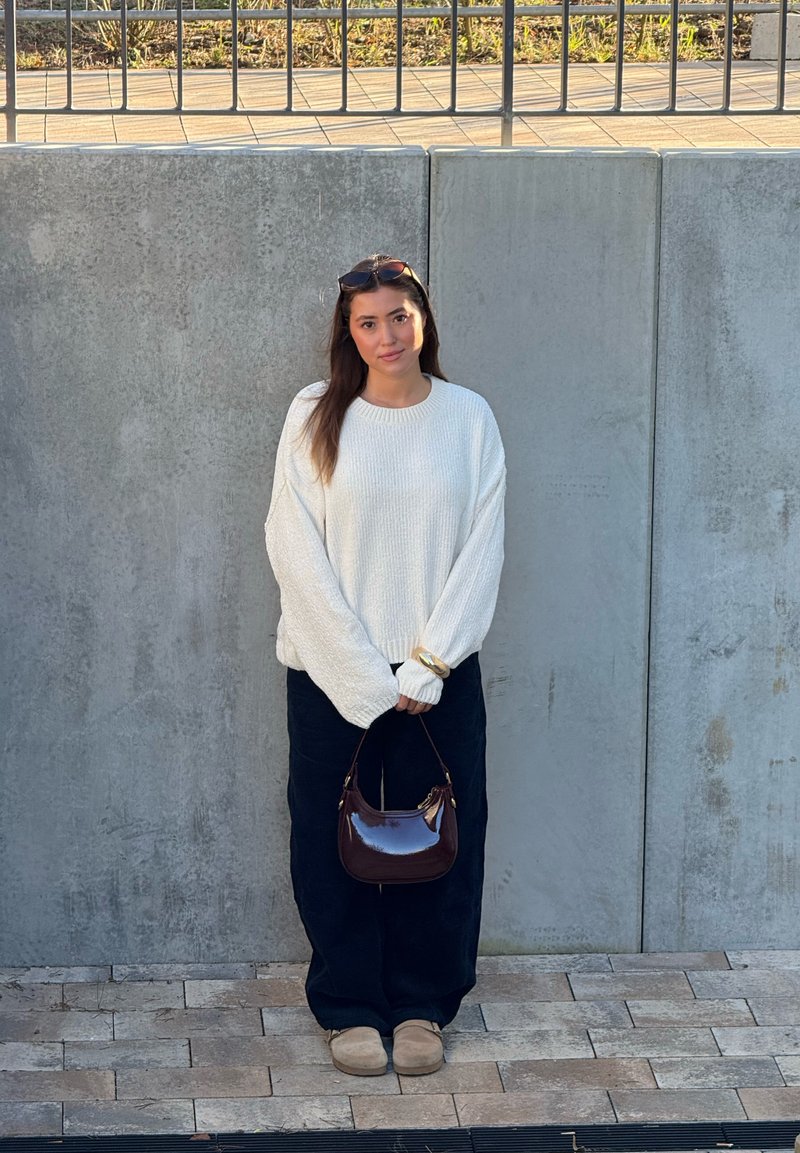 Femme en pull blanc, pantalon noir et chaussures beige, debout contre un mur en béton, tenant un petit sac à main marron et portant des lunettes de soleil sur la tête.