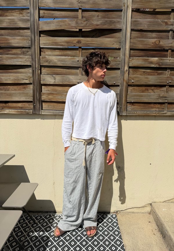 Jeune personne aux cheveux bouclés portant une chemise blanche à manches longues et un pantalon ample rayé, debout contre une clôture en bois au soleil.
