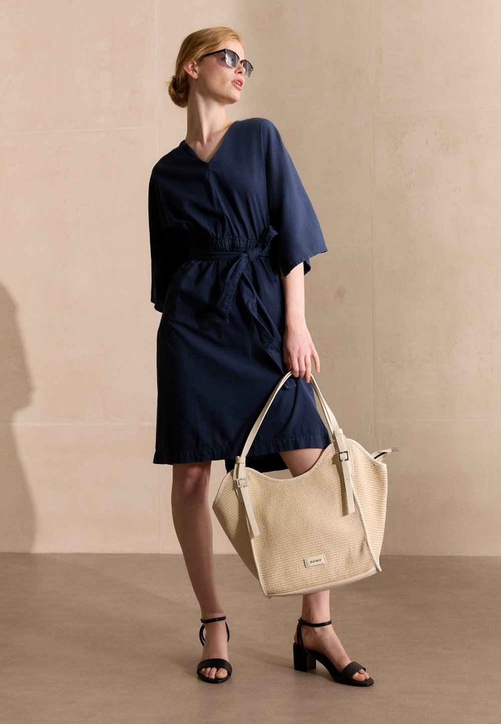 Marineblaues Wickelkleid mit Kordelzug an der Taille, kombiniert mit einer großen beigen geflochtenen Tote-Tasche und schwarzen Sandalen mit Blockabsatz.