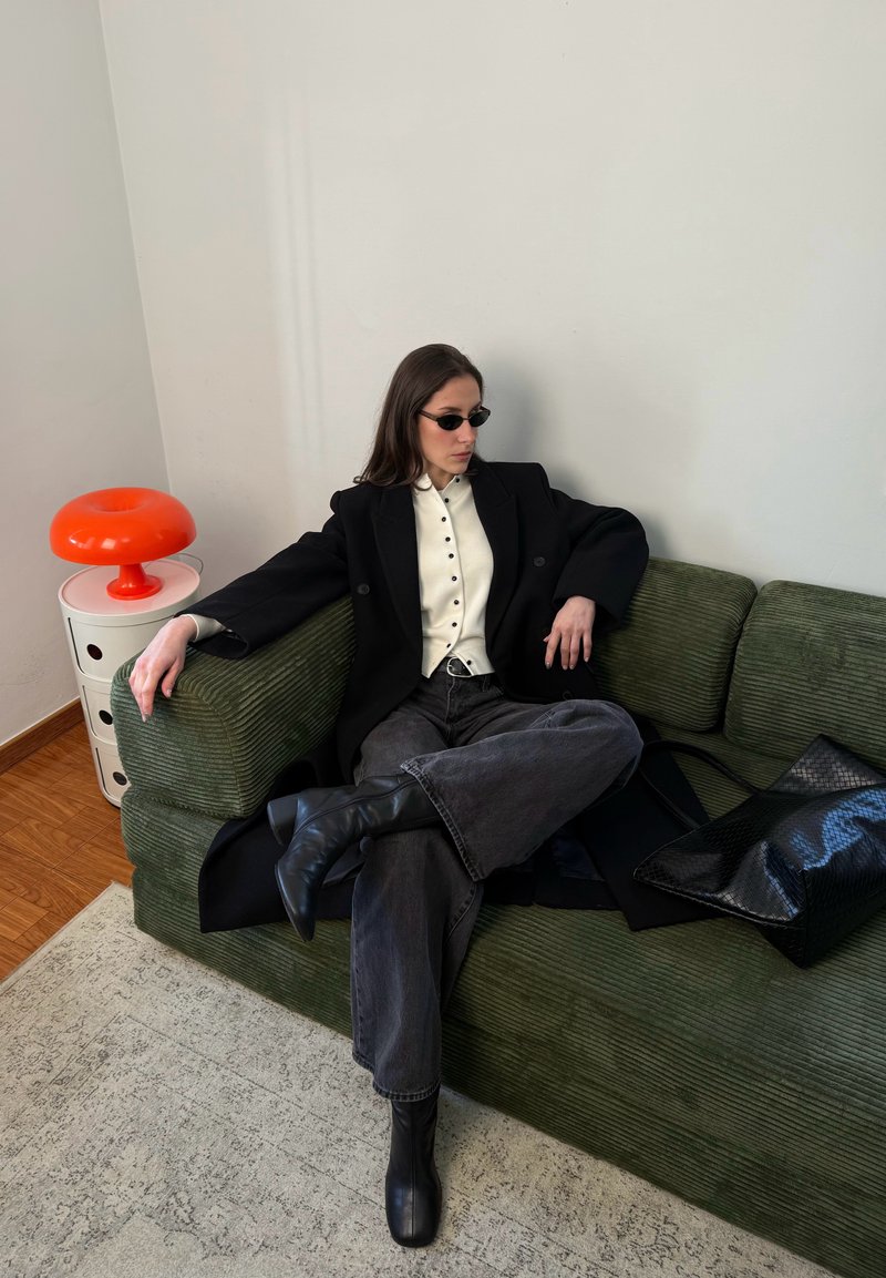 Femme portant un manteau noir, une chemise blanche à boutons, un jean foncé et des bottes, assise en tailleur sur un canapé vert avec un sac noir et une lampe orange à proximité.