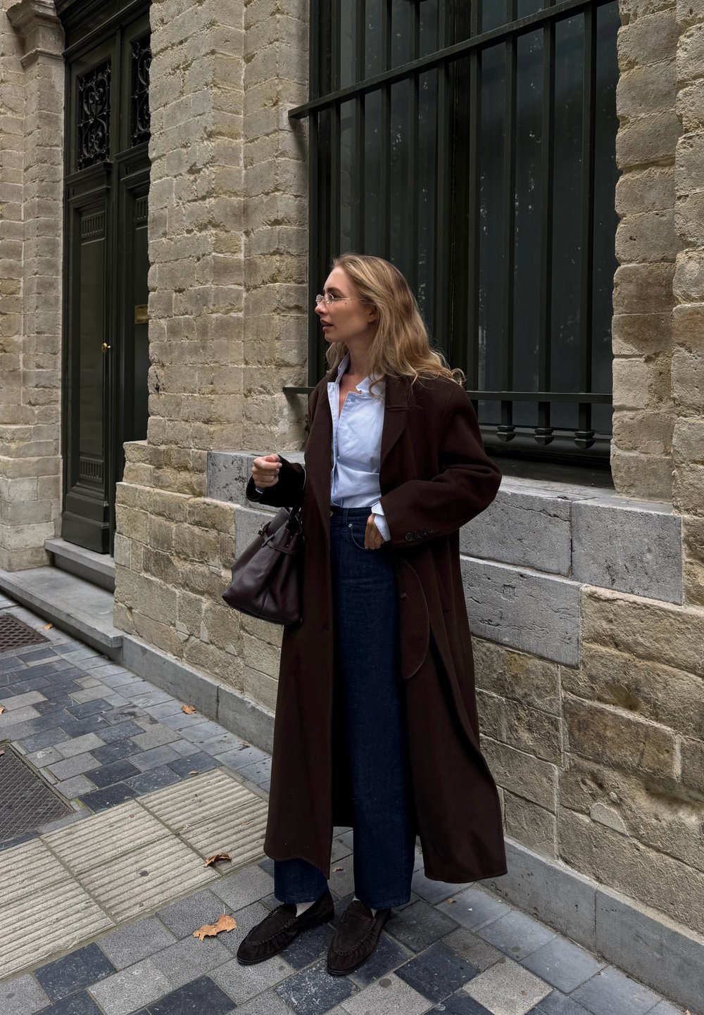Lungo cappotto marrone, camicia bianca, pantaloni in denim scuri a vita alta, mocassini scuri e borsa nera. In piedi contro un muro di pietra.