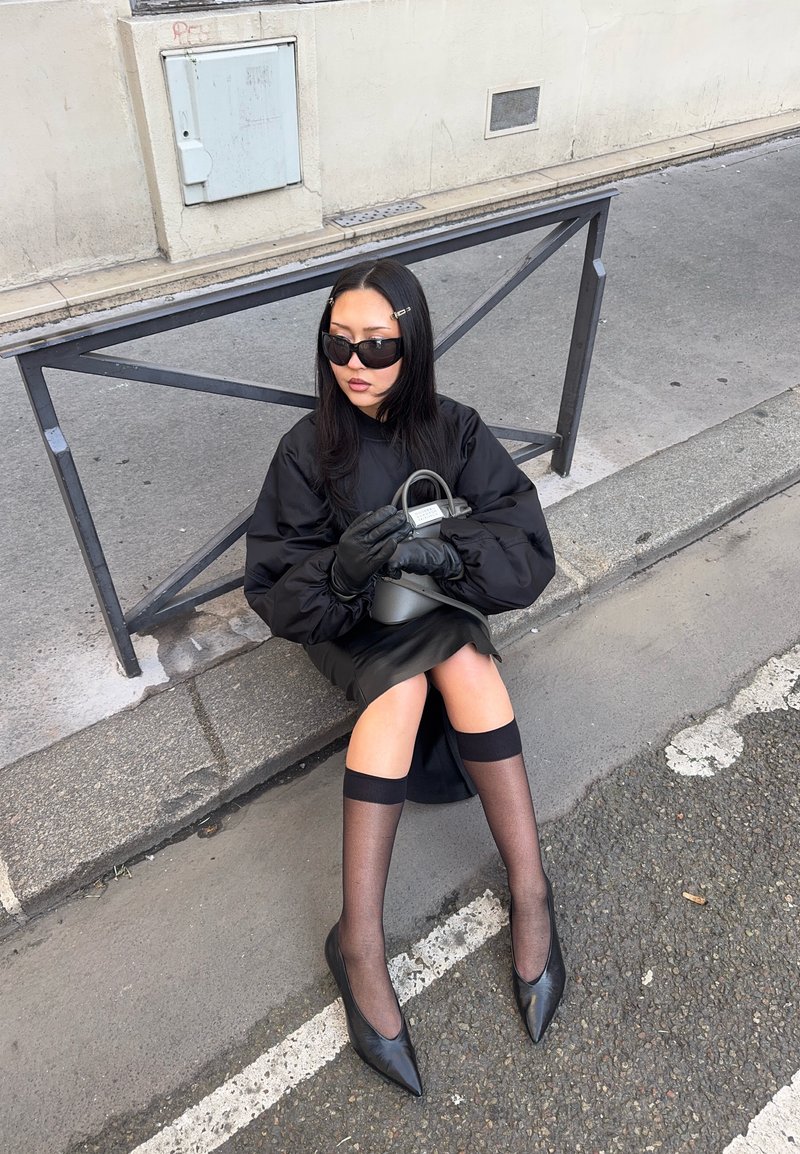 Femme portant des lunettes de soleil noires, des gants et une robe, assise sur un trottoir tenant un sac à main gris à côté d'une rambarde métallique dans une rue de la ville.