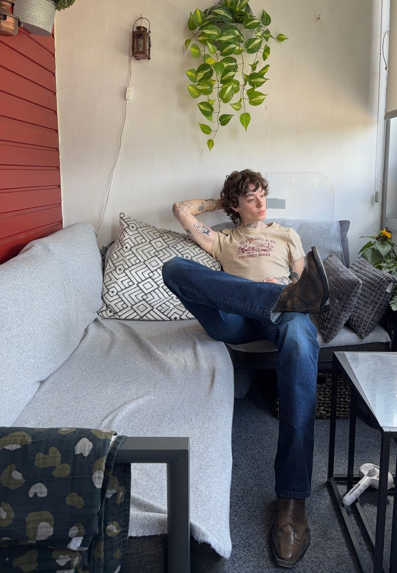 Personne avec des tatouages assise sur un canapé gris avec des coussins à motifs, portant une chemise beige et un jean, avec une plante verte suspendue au-dessus.