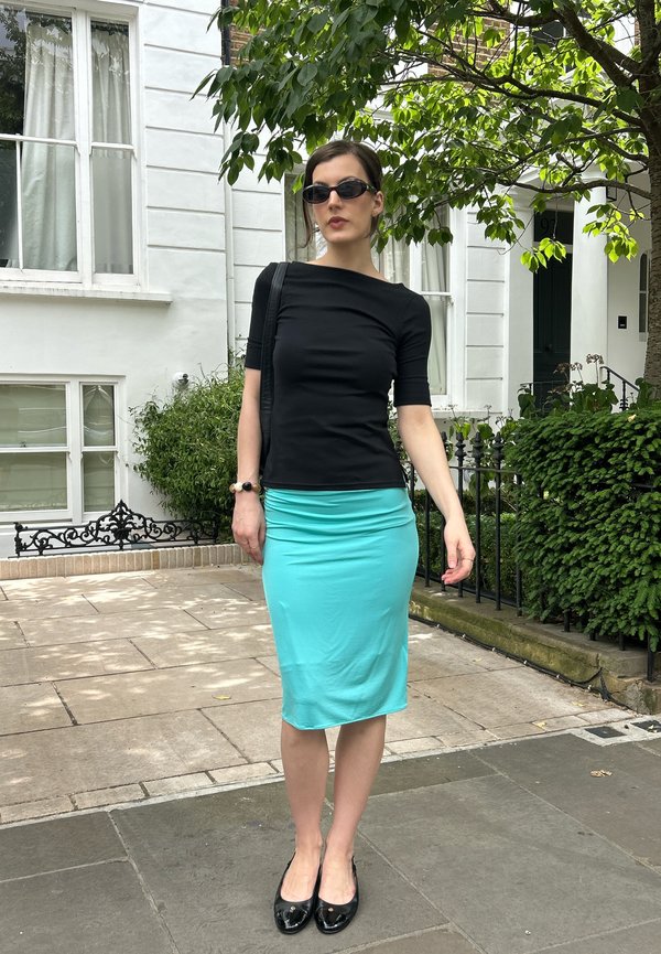 Woman wearing black top, turquoise skirt, black flats, and sunglasses stands on a sidewalk in front of a white building with greenery.