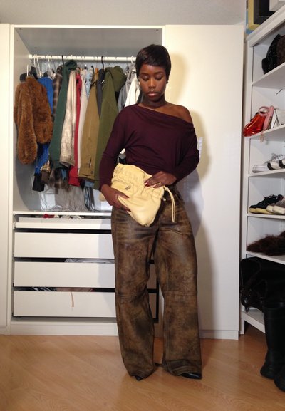 Una mujer con pantalones amplios marrones y una blusa burdeos sin hombros sostiene un clutch amarillo fruncido, de pie en una habitación con un armario abierto y estantes.